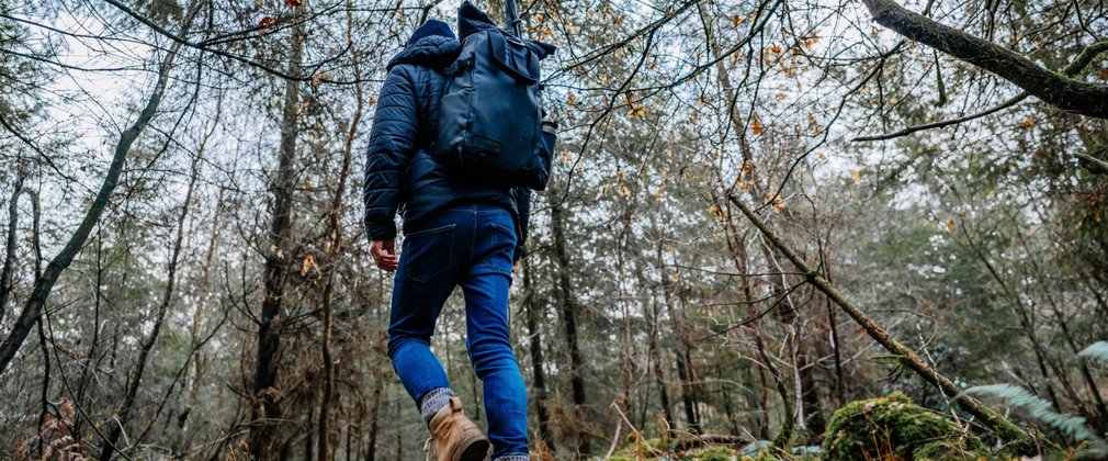 A man with his back to the camera, wearing walking boots, jeans, coat and hat, walking through the forest in winter.