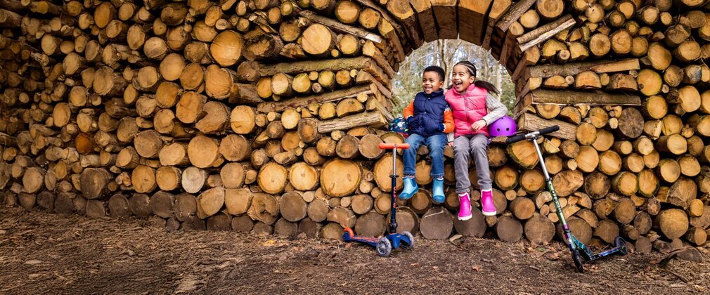 Two young children with scooters and helmets sit by a wall of stacked logs.