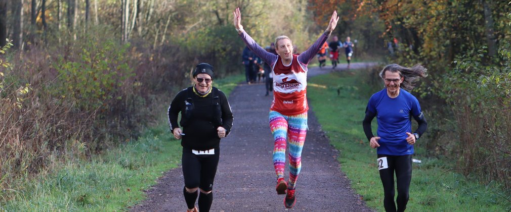 Two men and a woman running in the forest. The woman is jumping in the air.