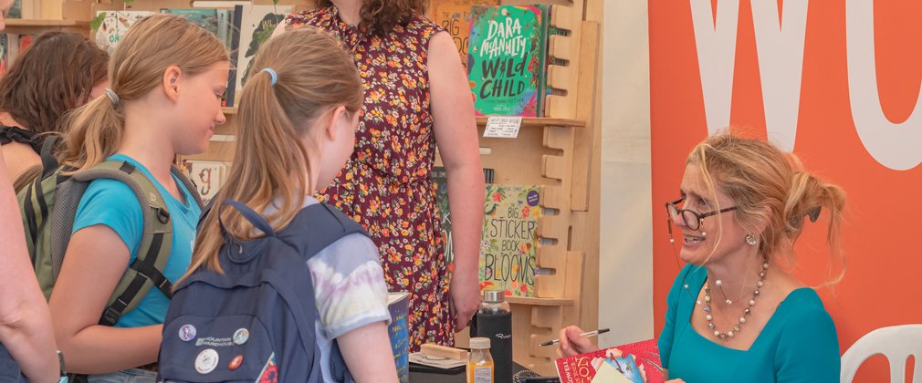 Two young girls talking across a table to a lady holding a book