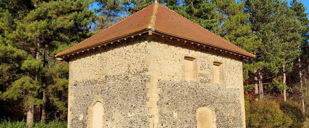 A square stone building with pitched roof shown in front of trees
