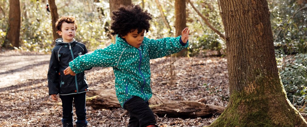 Two children in forest, one in coat and wellies is running over a dirt mound