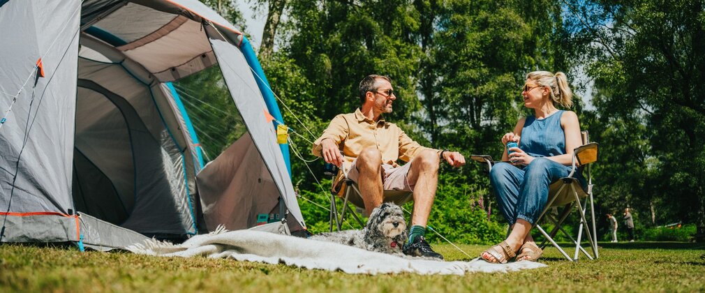 Two people sat on camp chairs next to a tent and dog lying on a blanket