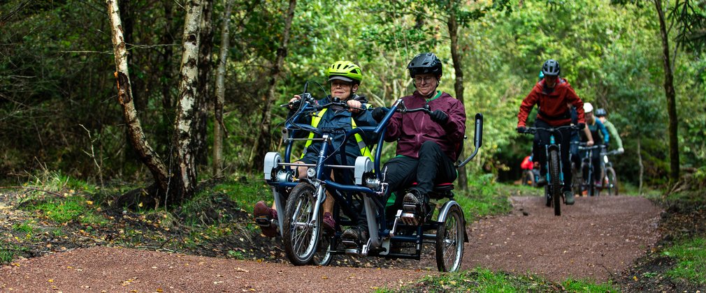 Two women riding a bike with two seats along a forest trail with other bike riders in the background