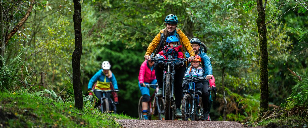 A group of riders cycling in a forest, led by a woman with a baby