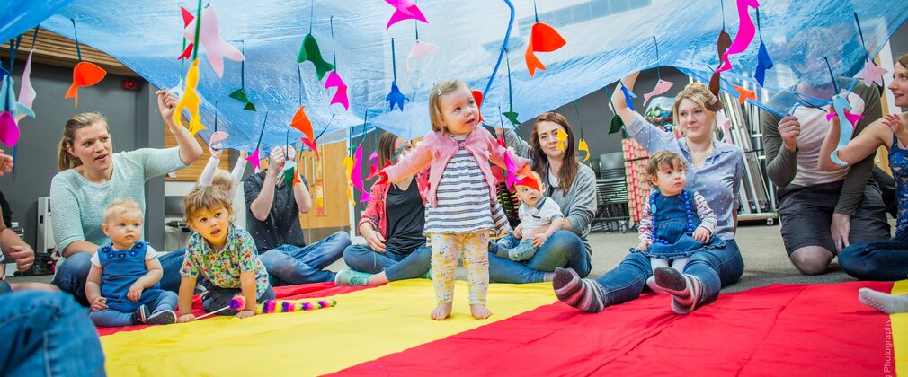 Image of a group of parents and small children playing under blue fabric with mobiles hanging