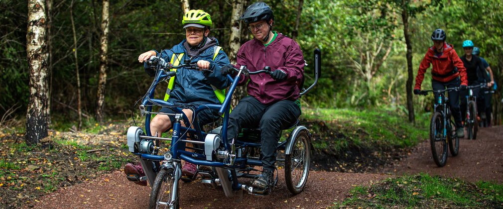 Two cyclists on a side-by-side adaptive bike, on a forest trail.
