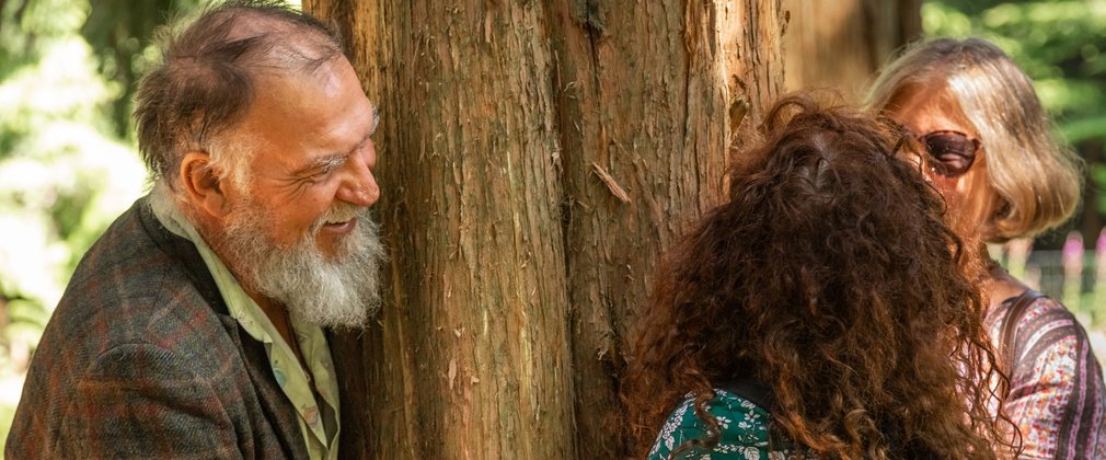 An older man and two older women smiling at each other and hugging a large tree trunk