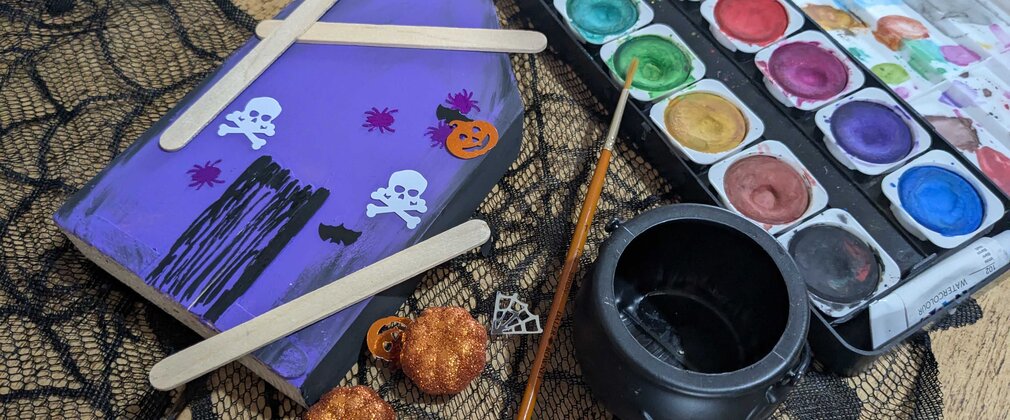 An arts and crafts image showing a watercolour paint set, paintbrush and a small painted wooden plaque of a haunted house