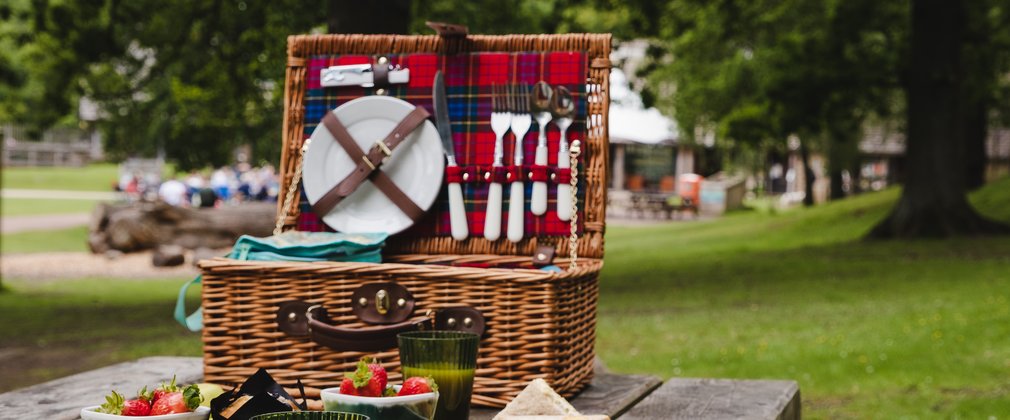 A wicker hamper with red tartan lining on a forest picnic table, shown with plates of food and drinks glasses.