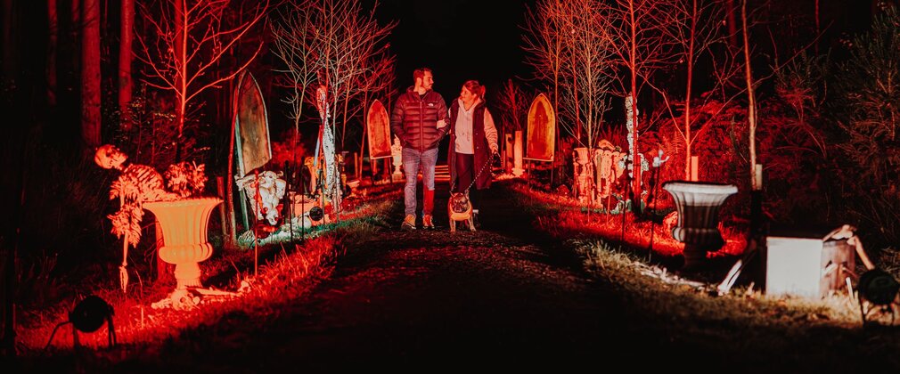 A couple walking a dog along a dark path bordered by spooky headstones.