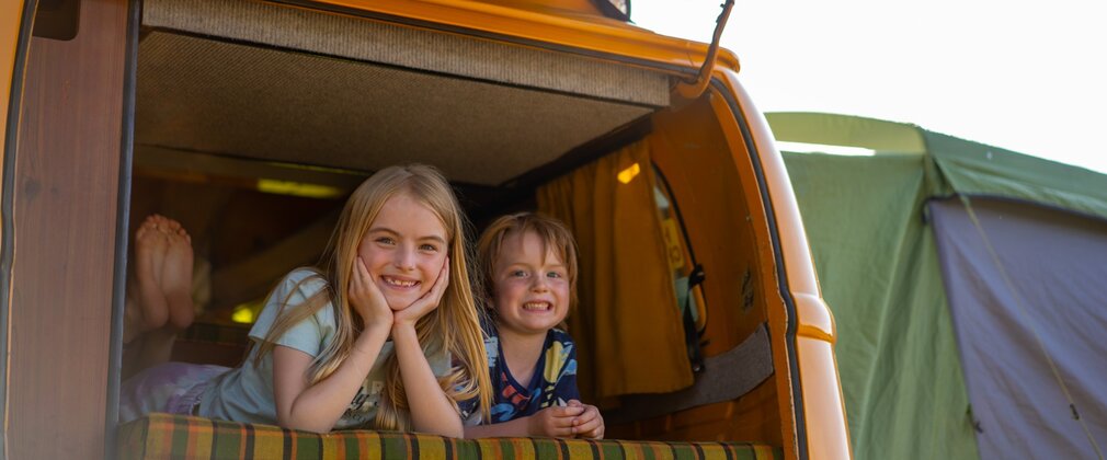 A boy and a girl smiling as they look out the back of an orange campervan