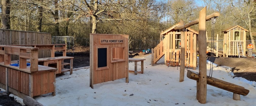 Sand play area featuring wooden play structures including houses, shop front and balance beam.