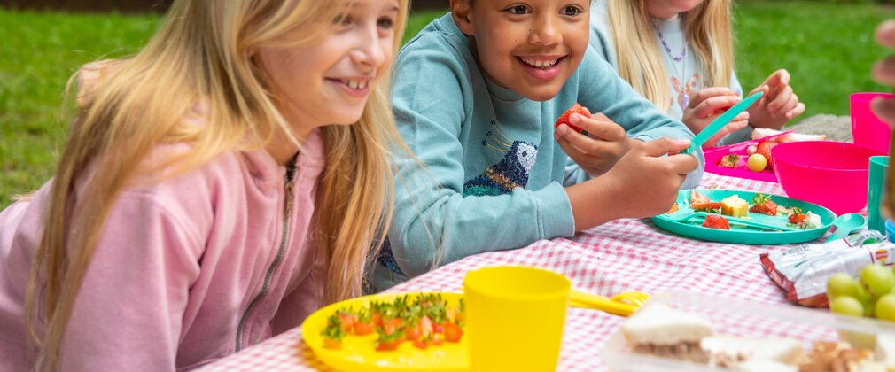 Three young girls with plates of fruit on a gingham-covered table outside.