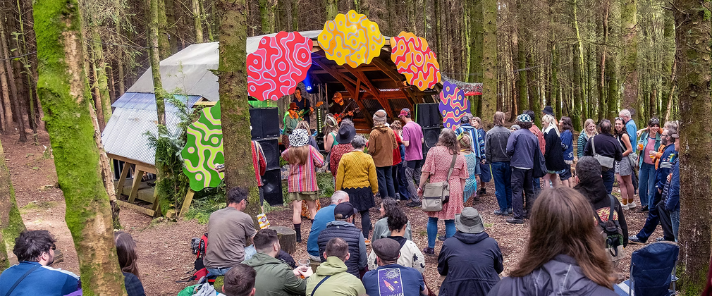 A group of people wihtin a woodland facing a stage with a band playing