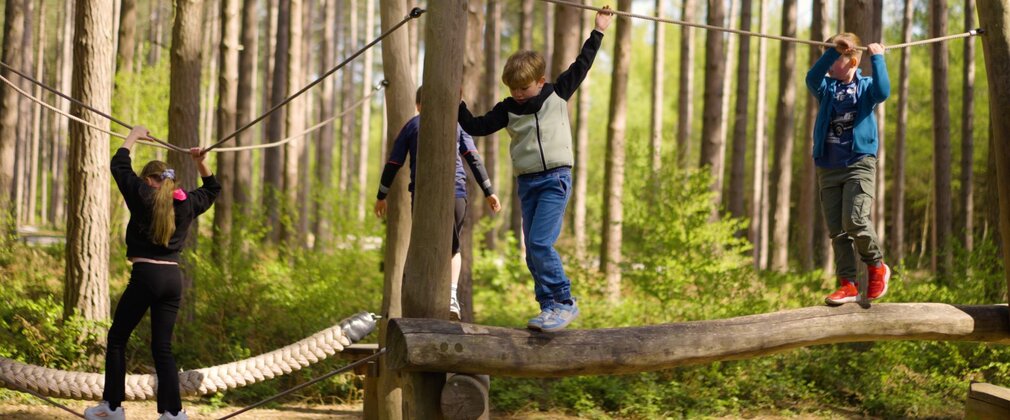 4 children on a wooden balance beam with a rope for them to hold onto.
