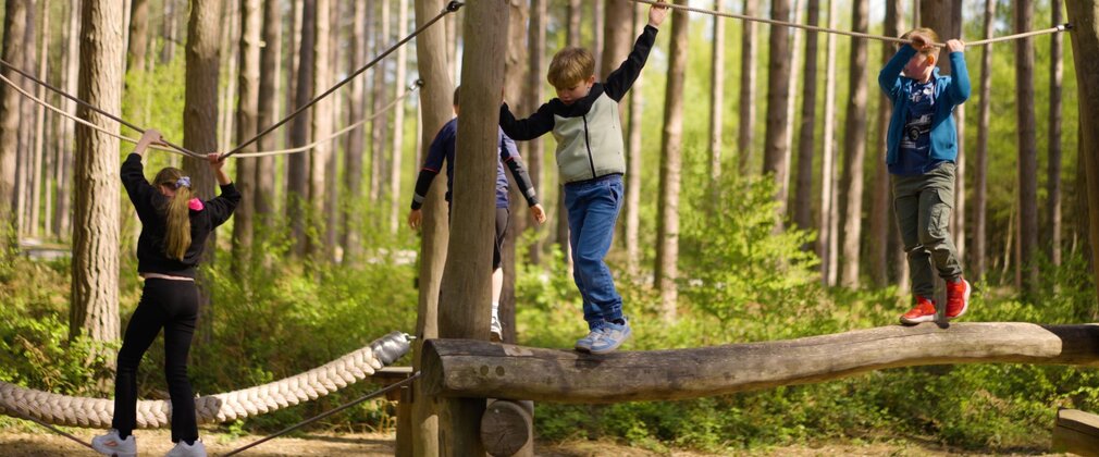 4 children on a wooden balance beam with a rope for them to hold onto.