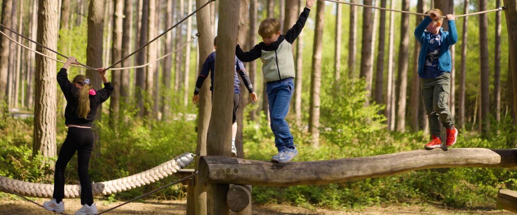 4 children on a wooden balance beam with a rope for them to hold onto.
