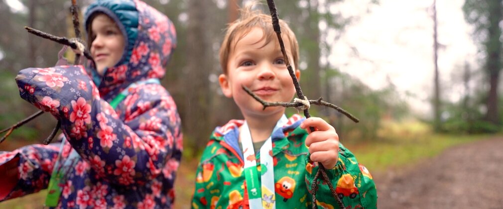 Two children on a forest path holding figures made from sticks.