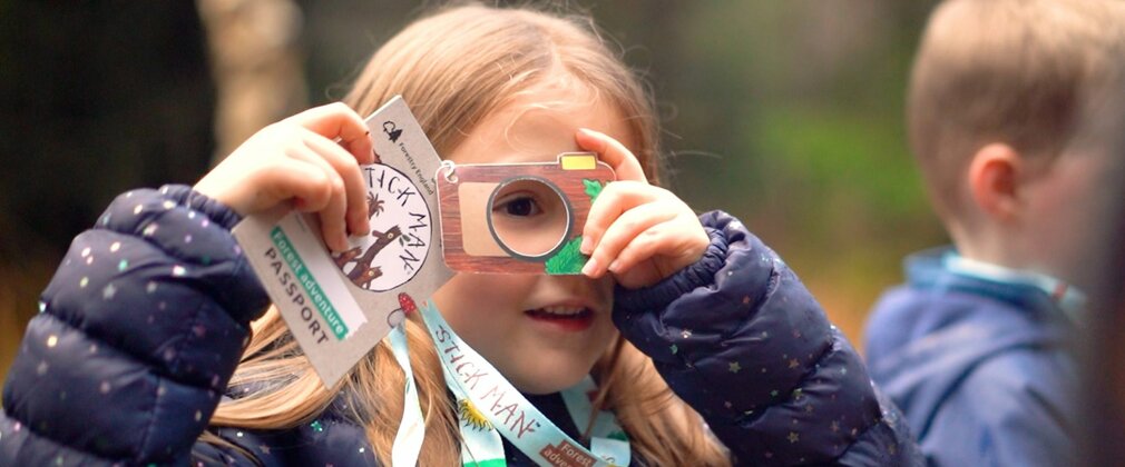 A girl in the forest looking through a viewfinder attached to a lanyard she's wearing.