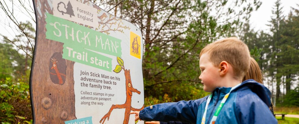 Two children in the forest pointing to a Stick Man branded panel