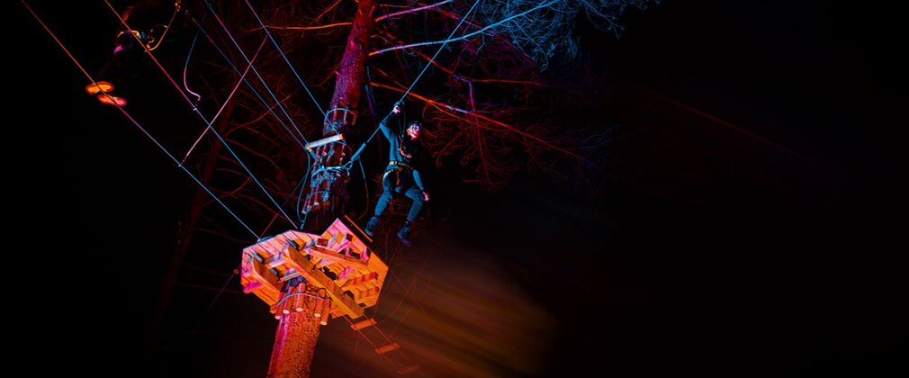 A person stepping off a platform from the top of a tree attached to a zip line, with a dark background and illuminated by orange light.