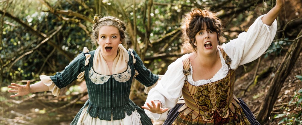 2 women dressed in medieval clothing look towards the audience with shocked expressions on their faces. Their hands stretch out as if they are signalling for hlp.