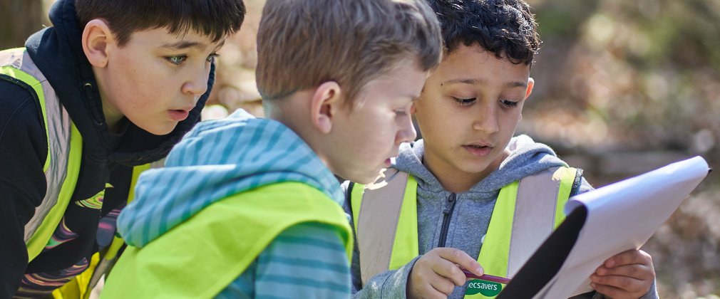 Three children looking at paper on a clipboard in a forest
