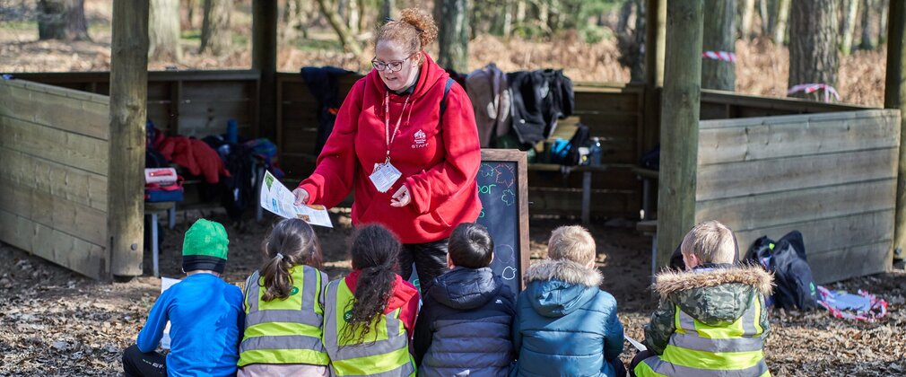 School pupils sit on a log gathered around their teacher leading an activity
