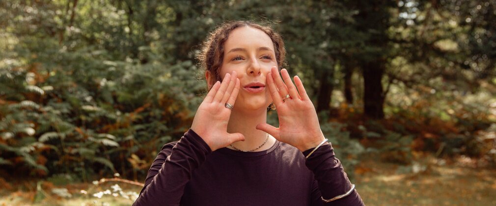 A woman in the forest holding her hands to either side of her mouth, in a calling gesture.