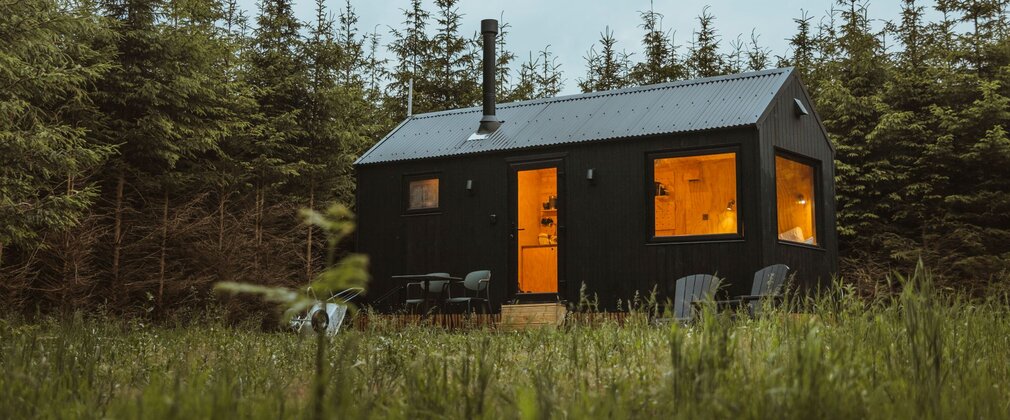 Small timber cabin with warm lights glowing through the windows, set in a grassy clearing and surrounded by dense evergreen forest.