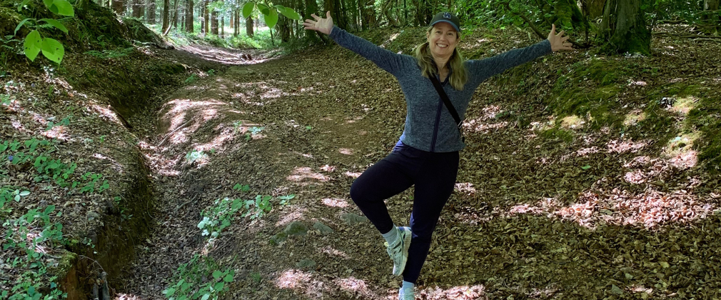 A woman stands with her arms wide open with one leg bent to place her foot by her calf in a yoga pose. She is surrounded by a forest landscape.