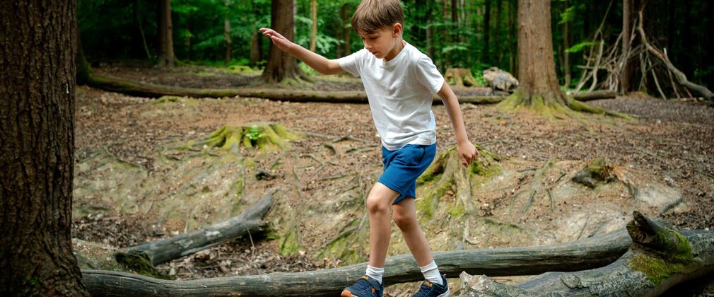 A boy in a white t-short and blue shorts balancing on a fallen log in a forest.