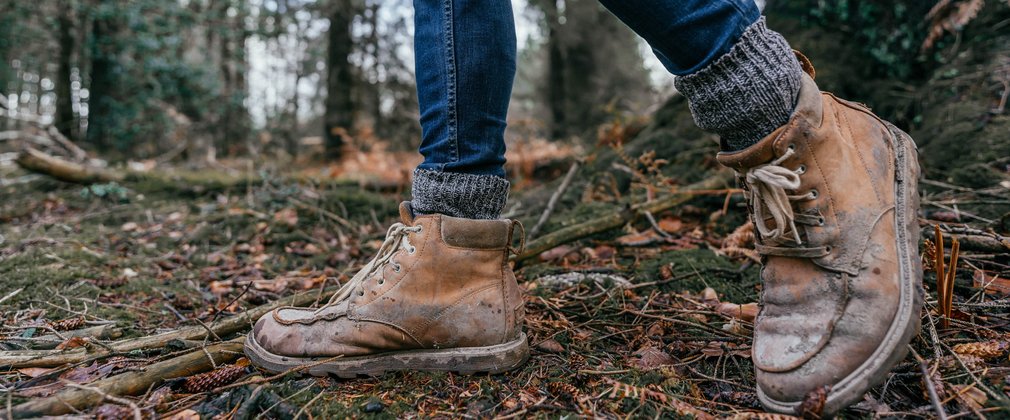 A pair of legs shown from the knees down, wearing well-worn walking boots, on a forest floor.