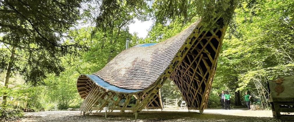 A wooden structure shaped like an armadillo shell made from long wooden laths and wooden shingles to create a Community Shelter