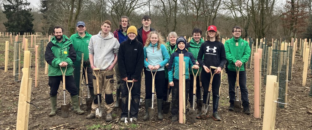 A group of adults and young people stand in a line for a photo. The young people hold spades in their hands, they have been planting trees in the area around them.