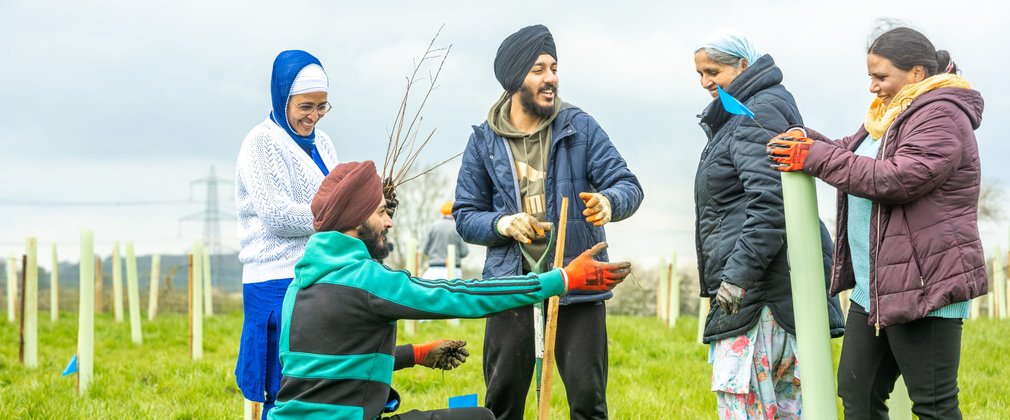A group of five adults at a tree planting event, one kneeling, one with a spade and one holding a tree sapling in protective material.