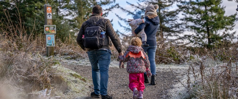With their backs to the camera, a family of two adults and two children walking on a forest path edged with frost. One of the children is wearing orange wings.