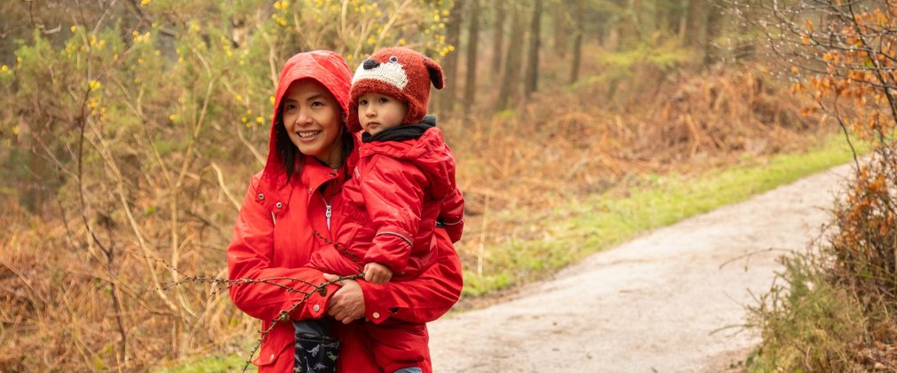 A woman in a red coat with hood up carries a child, wearing a red rain suit, along a forest path