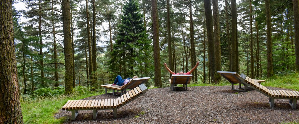 Two people lying on loungers under the trees. The man on the right has his arms outstretched.
