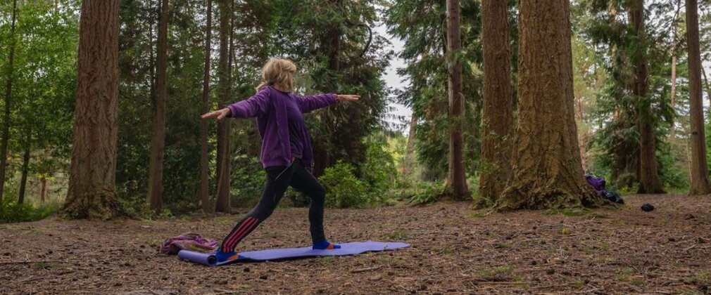 A woman doing a standing stretch on a yoga mat in a forest clearing.