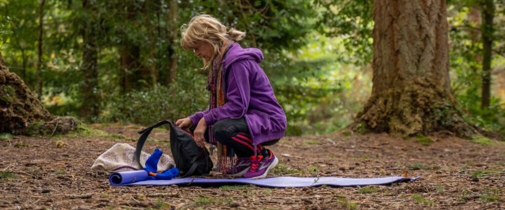 A woman crouches on a yoga mat looking at equipment in a forest clearing.