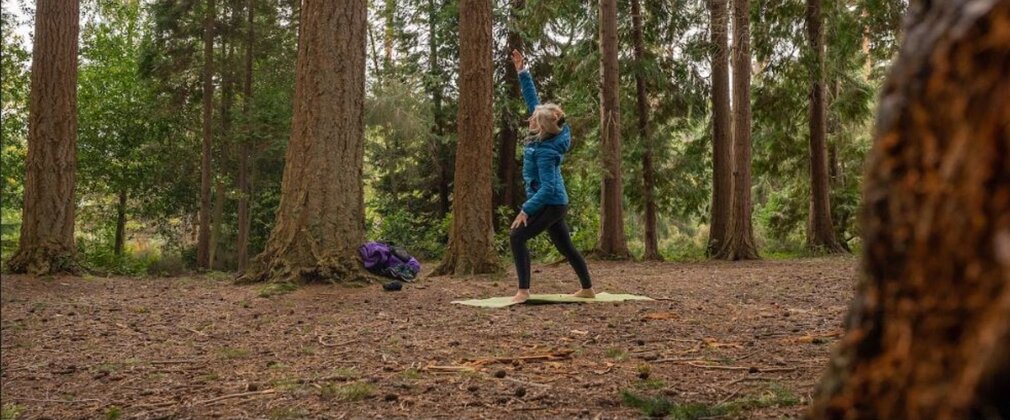 A woman doing a standing stretch on a yoga mat in a forest clearing.