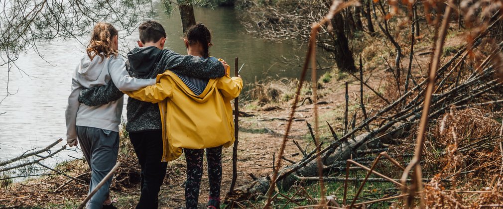 Three children walking together arm in arm, next to a lake