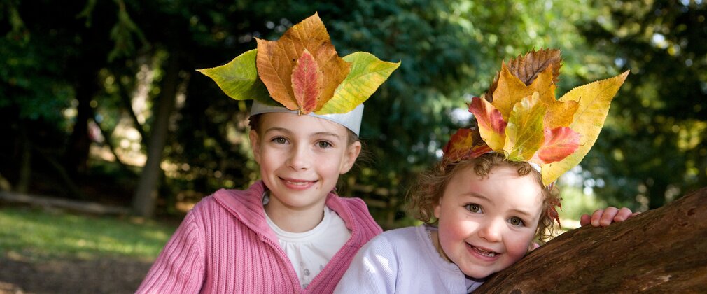 Two young girls, one in pink top and the other in white, wearing leaf crowns, standing at a tree branch and smiling.