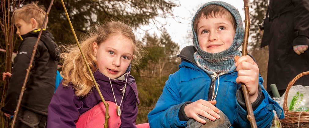 A young boy and girl face the camera, holding sticks as part of a bushcraft activity