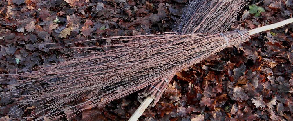 Two besom brooms made of wood sit on the floor one laid over the other.