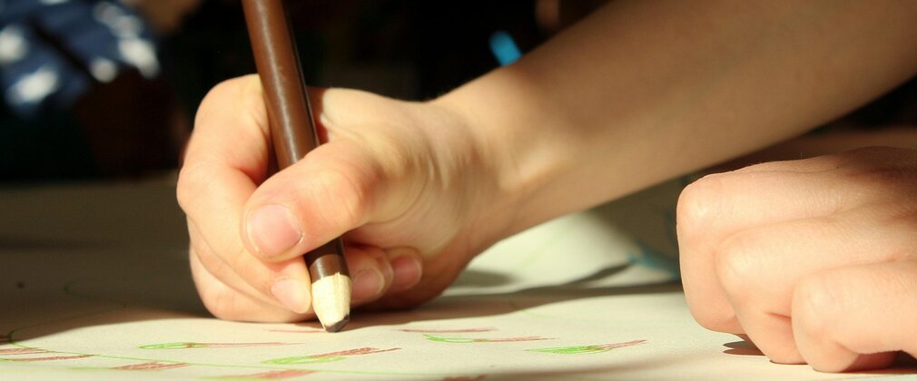 Close-up of a child's hand holding a coloured pencil, drawing on paper.