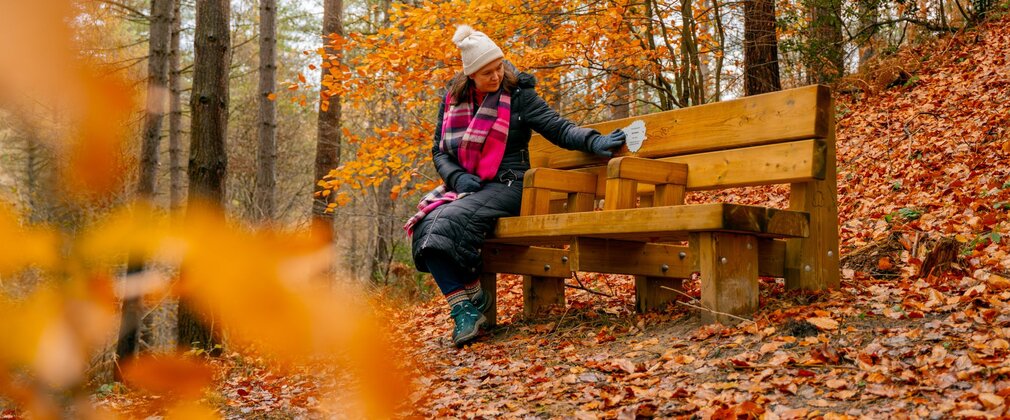 A woman sits on a wooden bench in the forest, looking at a memorial plaque.