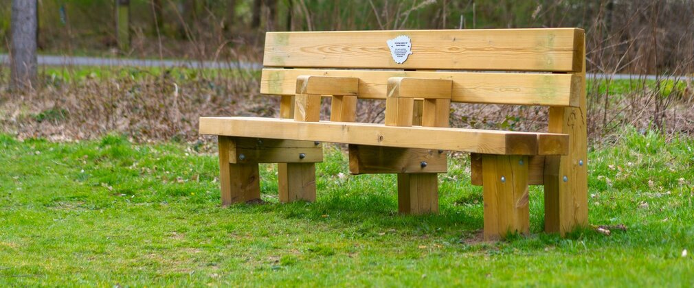 A wooden bench with arm rests and a leaf-shaped memorial plaque, situated in a forest.